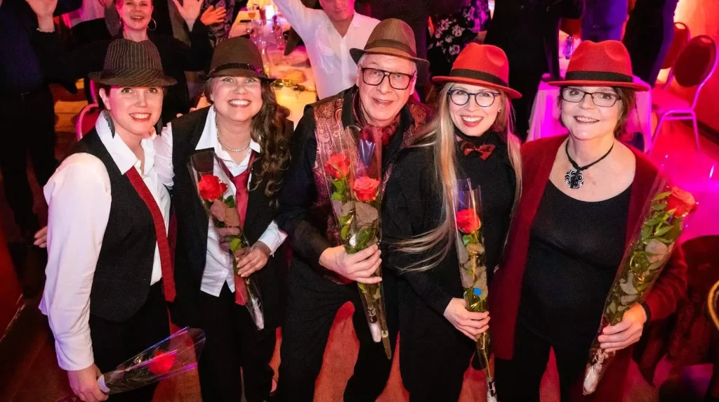 Sointu Ensemblen viisi hymyilevää esiintyjää seisoo lavan edessä pitäen pitkiä ruusuja kädessään. He ovat pukeutuneet tyylikkäisiin esiintymisasuihin, joihin kuuluu muun muassa hatut, liivit ja solmiot tai rusetit. Taustalla suuri yleisö juhlavassa salissa istuu pöytien äärellä ja heiluttaa iloisesti käsiään kohti kameraa. Valaistus on lämmin ja tunnelmallinen, punertavin ja violetin sävyin. Kuva huokuu yhteisöllisyyttä, iloa ja juhlan tuntua.
