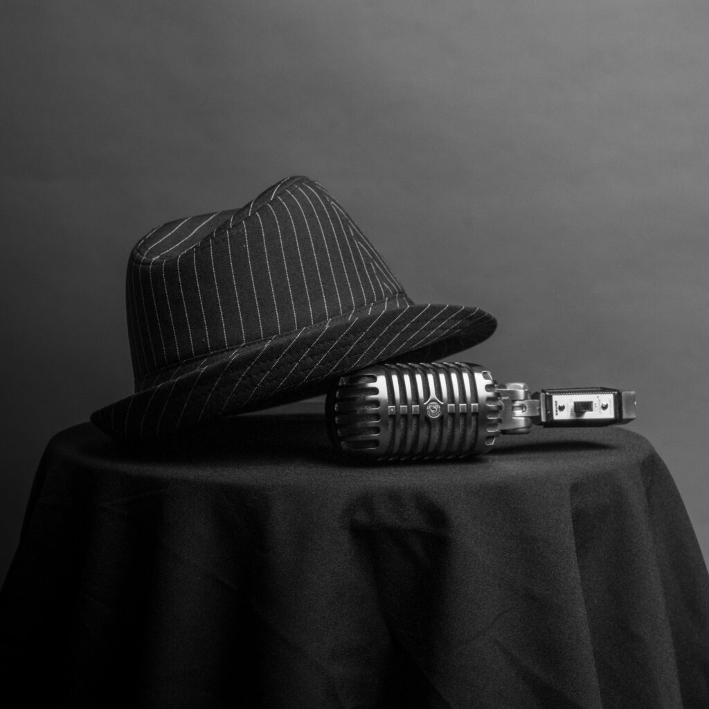 Mustavalkoinen still-kuva: raidallinen fedora-hattu ja vintage-mikrofoni mustalla pöydällä — Sointu Ensemble, Leonard Cohen tribute